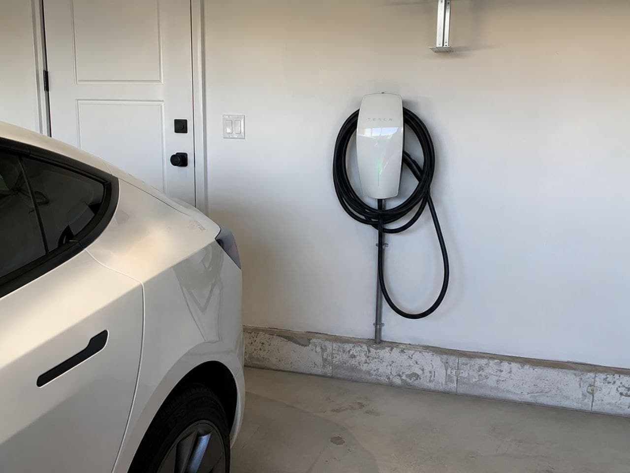 White Tesla parked in a garage next to a wall-mounted Tesla charger with coiled cable.